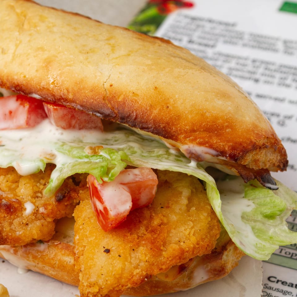 Close-up of a crispy chicken sandwich with lettuce, tomatoes, and white sauce on a baguette.