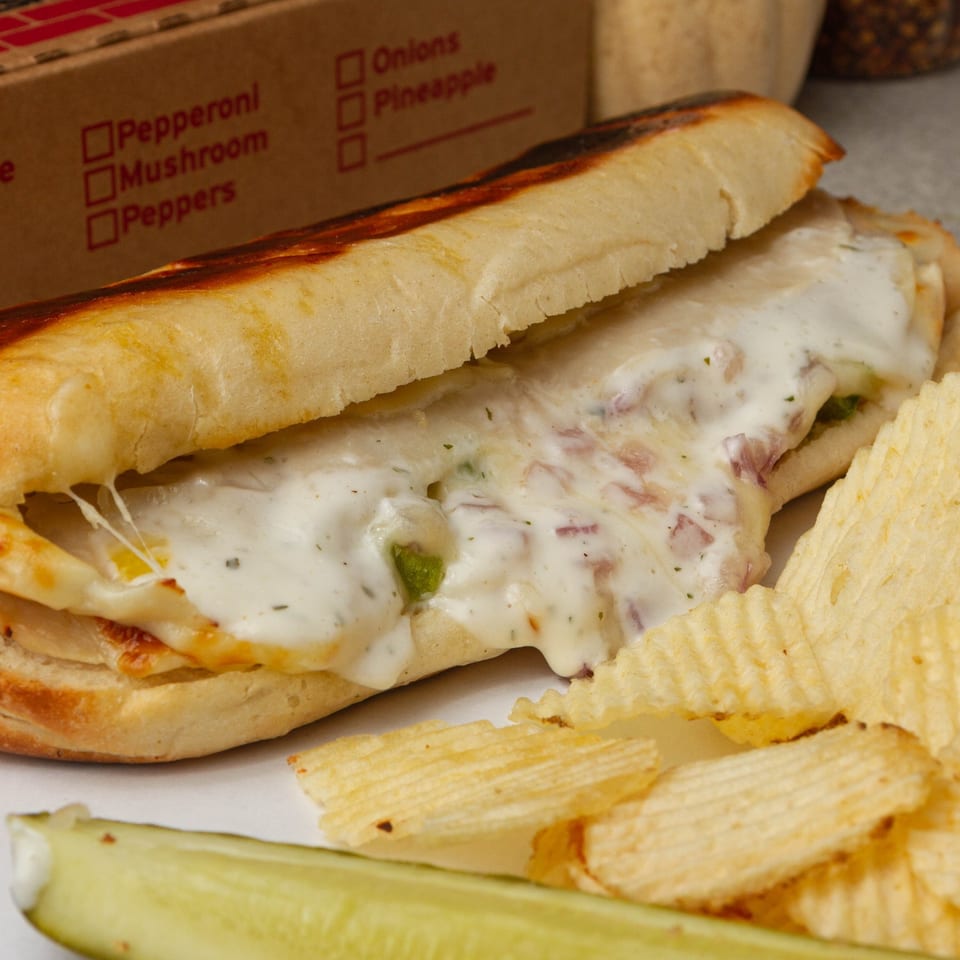 Cheesy Rounders Pizza sub sandwich with chips and a pickle spear, next to a pizza box.