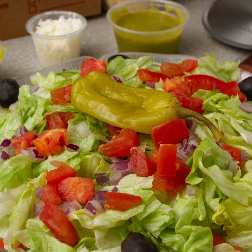 Fresh garden salad with lettuce, tomatoes, red onions, olives, and a pepperoncini, with dressing on the side.