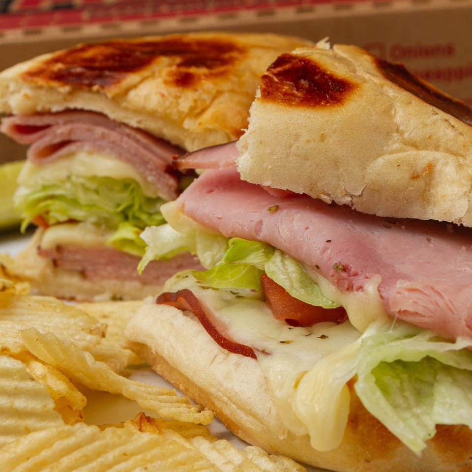 Two halves of a toasted sub sandwich with ham, melted cheese, lettuce, and tomatoes, served with potato chips.